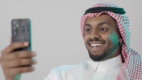 التواصل مع العائلة و الأصدقاء في أوقات الفراغ، إجراء مكالمة فيديو عبر الجوال , بورتريه مقرب لرجل عربي خليجي سعودي  يرتدي الثوب التقليدي و الشماغ  يمسك بيده الهاتف المحمول ,ايماءات السعادة و السرور ، خلفية رمادية
