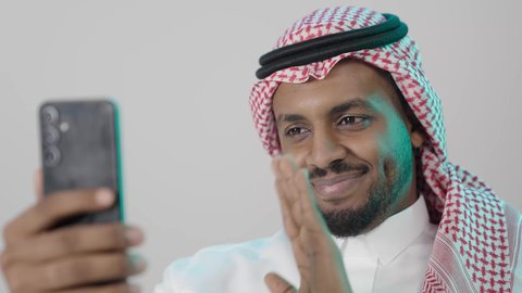 ايماءات يدين تدل على الترحيب، عرض بث مباشر على إحدى وسائل السوشل ميديا، التواصل مع العائلة و الأصدقاء في أوقات الفراغ، إجراء مكالمة فيديو عبر الجوال , بورتريه مقرب لرجل عربي خليجي سعودي  يرتدي الثوب التقليدي و الشماغ  يمسك بيده الهاتف المحمول ,تعابيرالسعادة و السرور ، خلفية رمادية