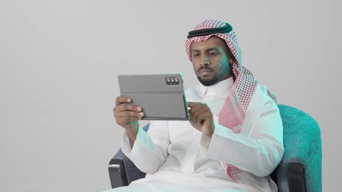إدارة المشاريع وتنظيم الأعمال عبر الإنترنت، تصفح مواقع و وسائل التواصل الاجتماعي، ايماءات التركيز والجدية ، تطبيقات وبرامج الأجهزة اللوحية، بورترية لرجل عربي خليجي سعودي يرتدي  الثوب الابيض و الشماغ يجلس على كرسي يستخدم جهاز التابلت ، خلفية رمادية