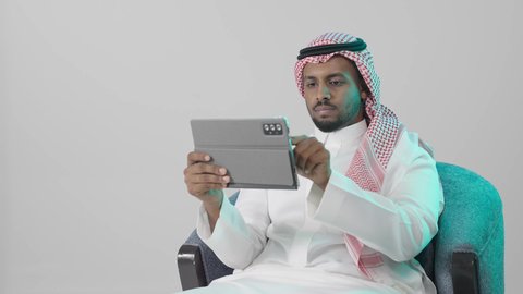 Monitoring performance and tasks remotely, browsing websites and social media, gestures of focus and seriousness, applications and programs for tablets, a portrait of a Saudi Arabian Gulf man wearing a white thobe and a ghutrah sitting on a chair using a tablet, gray background.