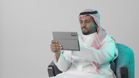 الإشراف على المهام والمشاريع أونلاين، تصفح مواقع و وسائل التواصل الاجتماعي، ايماءات التركيز والجدية ، تطبيقات وبرامج الأجهزة اللوحية، بورترية لرجل عربي خليجي سعودي يرتدي  الثوب الابيض و الشماغ يجلس على كرسي يستخدم جهاز التابلت ، خلفية رمادية