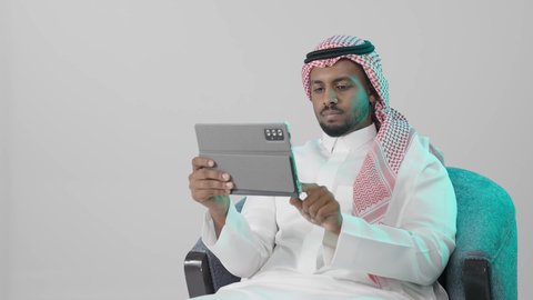 متابعة سير العمل والمهام عن بعد، تصفح مواقع و وسائل التواصل الاجتماعي، استخدام التكنولوجيا و التقنيات الحديثة، ايماءات التركيز والجدية ، تطبيقات وبرامج الأجهزة اللوحية، بورترية لرجل عربي خليجي سعودي يرتدي  الثوب الابيض و الشماغ يجلس على كرسي يستخدم جهاز التابلت ، خلفية رمادية