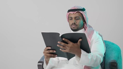 Independent learning and skill acquisition, carefully exploring the content of the book, a portrait of a young Arab Gulf Saudi man wearing a white thobe and a ghutrah sitting on a chair holding an educational book in his hand, gestures of focus and engagement, enjoying reading novels and stories, gray background.
