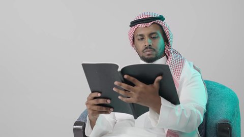 تطوير الذات من خلال التعلم الشخصي، استكشاف محتوى الكتاب بعناية ، بوترية لشاب عربي خليجي سعودي يرتدي الثوب الابيض و الشماغ يجلس على كرسي يحمل بيده كتاب تثقيفي ، إيماءات التركيز والاندماج ، الاستمتاع بقراءة الروايات والقصص، خلفية رمادية
