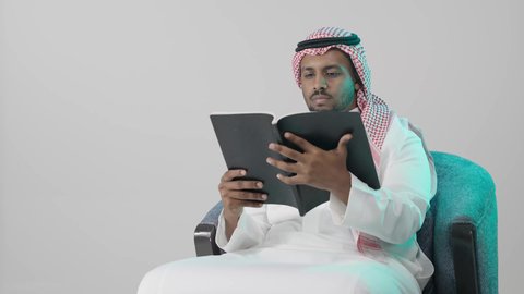 استكشاف محتوى الكتاب بعناية، ثقافة التعلم الذاتي ، بوترية لشاب عربي خليجي سعودي يرتدي الثوب الابيض و الشماغ يجلس على كرسي يحمل بيده كتاب تثقيفي ، إيماءات التركيز والاندماج ، الاستمتاع بقراءة الروايات والقصص، خلفية رمادية