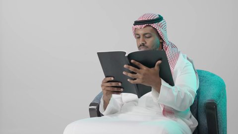 تحليل المعلومات من الكتب بعناية، ثقافة التعلم الذاتي ، بوترية لشاب عربي خليجي سعودي يرتدي الثوب الابيض و الشماغ يجلس على كرسي يحمل بيده كتاب تثقيفي ، إيماءات التركيز والاندماج ، الاستمتاع بقراءة الروايات والقصص، خلفية رمادية