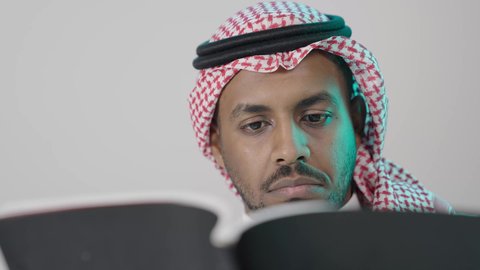 A careful reading of the book, the culture of self-learning, a close portrait of a young Arab Gulf Saudi man wearing a white thobe and a shemagh holding an educational book in his hand, gestures of focus and engagement, enjoying reading novels and stories, gray background.