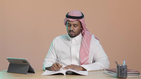 المذاكرة للاختبارات الجامعية، حل الواجبات الجامعية، بورتريه لطالب جامعي عربي خليجي سعودي يرتدي الثوب الابيض و الشماغ يجلس على مكتب يقوم بتدوين الملاحظات و قراءة الكتب ، مراجعة الدروس اليومية ، التعليم الجامعي في المملكة العربية السعودية، خلفية بني