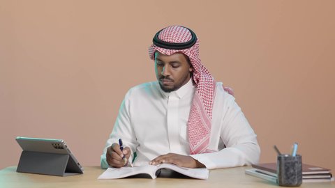 حل الواجبات الجامعية، بورتريه لطالب جامعي عربي خليجي سعودي يرتدي الثوب الابيض و الشماغ يجلس على مكتب يقوم بتدوين الملاحظات و قراءة الكتب ،المذاكرة للاختبارات الجامعية ، مراجعة الدروس اليومية ، التعليم الجامعي في المملكة العربية السعودية، خلفية بني