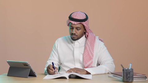 بورتريه لطالب جامعي عربي خليجي سعودي يرتدي الثوب الابيض و الشماغ يجلس على مكتب يقوم بتدوين الملاحظات و قراءة الكتب ، حل الواجبات الجامعية ،المذاكرة للاختبارات الجامعية ، مراجعة الدروس اليومية ، التعليم الجامعي في المملكة العربية السعودية، خلفية بني