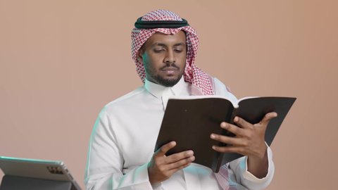 قراءة الكتاب بشكل مركز، ثقافة التعلم الذاتي ، بوترية لشاب عربي خليجي سعودي يرتدي الثوب الابيض و الشماغ يحمل بيده كتاب تثقيفي ، إيماءات التركيز والاندماج ، الاستمتاع بقراءة الروايات والقصص، خلفية بنية