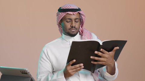 Diving into the content of books, the culture of self-learning, a portrait of a young Arab Gulf Saudi man wearing a white thobe and a shemagh holding an educational book in his hand, gestures of focus and immersion, enjoying reading novels and stories, brown background.
