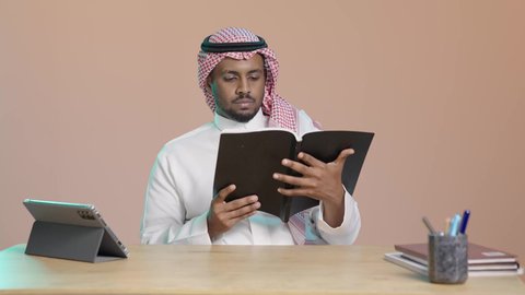 Studying books carefully, the culture of self-learning, a close portrait of a young Arab Gulf Saudi man wearing a white thobe and a shemagh sitting at a desk holding an educational book, gestures of focus and engagement, enjoying reading novels and stories, brown background.