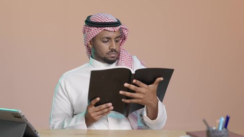 Reading books attentively, the culture of self-learning, a close portrait of a young Arab Gulf Saudi man wearing a white thobe and a shemagh sitting at a desk holding an educational book, gestures of focus and engagement, enjoying reading novels and stories, brown background.