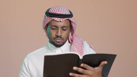 ثقافة التعلم الذاتي، قراءة الكتب بتمعن ، بوترية مقرب لشاب عربي خليجي سعودي يرتدي الثوب الابيض و الشماغ يحمل بيده كتاب تثقيفي ، إيماءات التركيز والاندماج ، الاستمتاع بقراءة الروايات والقصص، خلفية بنية