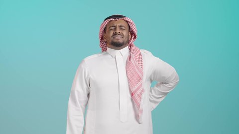 التعب والإرهاق والإحساس بالمرض، بورتريه لرجل عربي خليجي سعةدي يرتدي الثوب الابيض و الشماغ يضع يده على ظهره، آلام الظهر والعامود الفقري، إيماءات وجه ويدين تدل على الوجع والألم، الحاجة إلى العلاج، خلفية خضراء