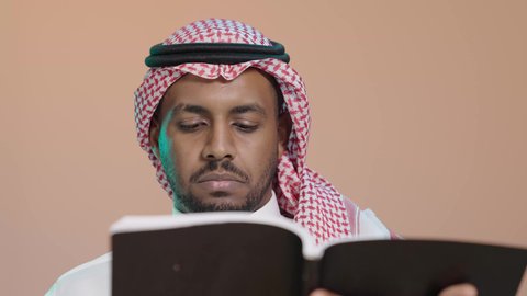 ثقافة التعلم الذاتي، قراءة الكتب بتمعن ، بوترية مقرب لشاب عربي خليجي سعودي يرتدي الثوب الابيض و الشماغ يحمل بيده كتاب تثقيفي ، إيماءات التركيز والاندماج ، الاستمتاع بقراءة الروايات والقصص، خلفية بنية