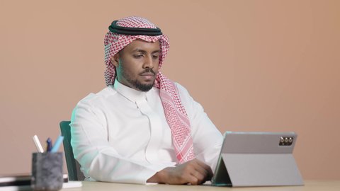 تصفح مواقع و وسائل التواصل الاجتماعي، مفهوم إدارة المشاريع وانجاز المهام عن بعد، استخدام التكنولوجيا و التقنيات الحديثة، ايماءات التركيز والجدية ، تطبيقات وبرامج الأجهزة اللوحية، بورترية لرجل عربي خليجي سعودي يرتدي  الثوب الابيض و الشماغ يجلس على مكتب يستخدم جهاز التابلت ، خلفية بني
