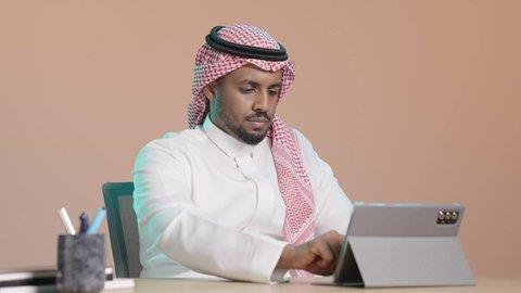 مفهوم إدارة المشاريع وانجاز المهام عن بعد، استخدام التكنولوجيا و التقنيات الحديثة، ايماءات التركيز والجدية ، تطبيقات وبرامج الأجهزة اللوحية، بورترية لرجل عربي خليجي سعودي يرتدي  الثوب الابيض و الشماغ يجلس على مكتب يستخدم جهاز التابلت ، تصفح مواقع و وسائل التواصل الاجتماعي ، خلفية بني