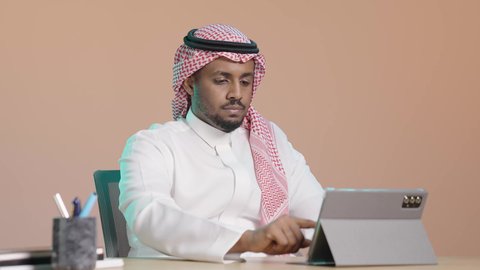 The use of technology and modern techniques, gestures of focus and seriousness, applications and programs for tablets, the concept of project management and completing tasks remotely, a portrait of a Saudi Arabian Gulf man wearing a white thobe and a ghutrah sitting at a desk using a tablet, browsing websites and social media, brown background.