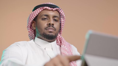 تطبيقات وبرامج الأجهزة اللوحية، ايماءات التركيز والجدية ، استخدام التكنولوجيا و التقنيات الحديثة، مفهوم إدارة المشاريع وانجاز المهام عن بعد، بورترية لرجل عربي خليجي سعودي يرتدي  الثوب الابيض و الشماغ يحمل جهاز التابلت ، تصفح مواقع و وسائل التواصل الاجتماعي ، خلفية بني
