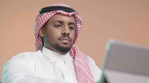 استخدام جهاز تقني حديث متطور، أداء المهام من المنزل، العمل عن بعد، بورترية مقرب لرجل عربي خليجي سعودي يرتدي الثوب الابيض و الشماغ يعمل على جهاز التابلت، دمج التقنية والتكنولوجيا في الحياة اليومية، خلفية بني