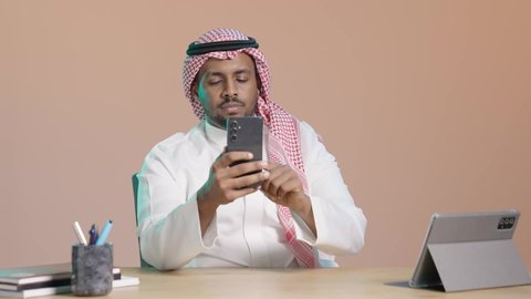التواصل الاجتماعي عبر الإنترنت، تصفح التطبيقات و البرامج، العمل عن بعد، استخدام جهاز تقني حديث ومتطور، بورترية مقرب لرجل عربي خليجي سعودي يرتدي الثوب الابيض و الشماغ يجلس على مكتب و يمسك بيده الهاتف المحمول، خلفية بني