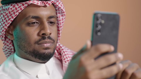 مشاركة الأخبار مع الأهل والأصدقاء، تصفح التطبيقات وبرامج التواصل الاجتماعي، العمل عن بعد، استخدام جهاز تقني حديث ومتطور، بورترية مقرب لرجل عربي خليجي سعودي يرتدي الثوب الابيض و الشماغ يمسك بيده الهاتف المحمول، خلفية  بني