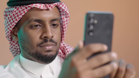 Chatting and exchanging conversations online, browsing applications and social media programs, working remotely, using a modern and advanced technical device, a close-up portrait of a Saudi Arabian Gulf man wearing a white thobe and a shemagh holding a mobile phone in his hand, brown background.
