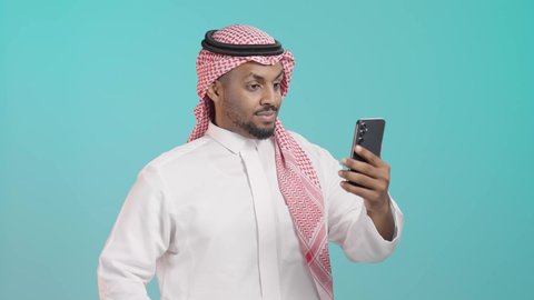 رفع الكف للأعلى بايماءات التحية والسلام، استخدام الأجهزة التقنية الحديثة، التواصل مع الأهل والاصدقاء، اجراء مكالمة فيديو عبر الهاتف المحمول، بورترية لرجل عربي خليجي سعودي يرتدي الثوب التقليدي والشماغ يقوم ببث مباشر عبر الموبايل، خلفية خضراء