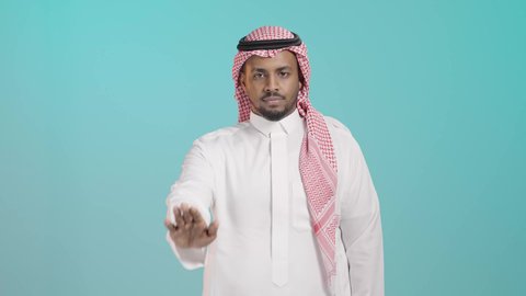 The concept of rejection, refusal, and discomfort, a request to stop and not approach, a portrait of a young Arab Gulf Saudi man wearing a white thobe and a shemagh looking at the camera, extending his hands forward with his palm raised in a gesture of prohibiting an action, green background.