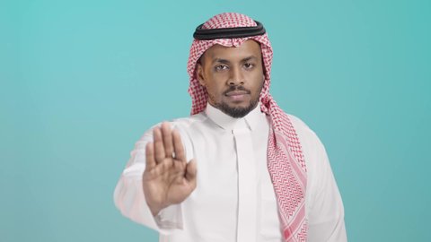 مفهوم الرفض والامتناع والانزعاج, طلب التوقف وعدم الاقتراب, بورتريه  لشاب عربي خليجي سعودي يرتدي الثوب الابيض و الشماغ ينظر الى الكاميرا يمد يديه نحو الأمام مع رفع الكف بتعابير النهي عن فعل امر ما, خلفية خضراء