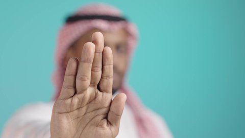 Request to stop and not approach, the concept of rejection, refusal, and discomfort, a close-up portrait of a young Arab Gulf Saudi man wearing a white thobe and a shemagh looking at the camera extending his hands forward with his palm raised in a gesture of prohibiting an action, green background.