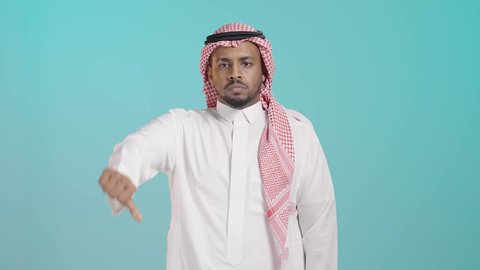 حركة يد وايماءات وجه تدل على عدم الإعجاب والموافقة، بورتريه لرجل عربي خليجي سعودي يرتدي الثوب الابيض و الشماغ تظهر عليه علامات عدم القبول، الإشارة بإصبع الإبهام للاسفل دليل الرفض، خلفية خضراء