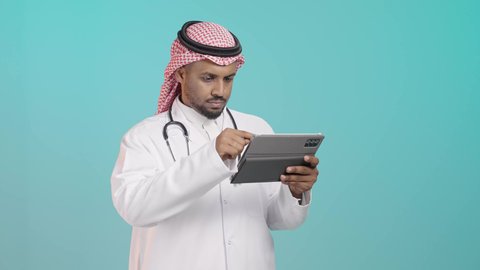 متابعة المرضى عبر التطبيقات الطبية، مفهوم الطب والرعاية الصحية الإلكترونية المنزلية، بورتريه لطبيب عربي خليجي سعودي يرتدي المعطف الطبي الأبيض و السماعات الطبية و الشماع يقوم بأستخدام جهاز التابلت لإعداد التقارير الطبية، خلفية خضراء