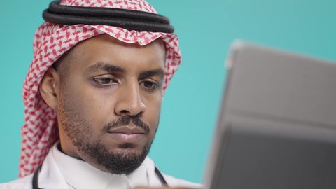 دراسة و معاينة الحالة المرضية للمرضى، تقديم الاستشارات الطبية عن بعد، بورتريه مقرب لطبيب عربي خليجي سعودي يرتدي المعطف الطبي الأبيض و السماعات الطبية و الشماع يقوم بأستخدام جهاز التابلت لإعداد التقارير الطبية، مفهوم الطب والرعاية الصحية الإلكترونية المنزلية، خلفية خضراء