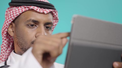 تقديم الاستشارات الطبية عن بعد، بورتريه مقرب لطبيب عربي خليجي سعودي يرتدي المعطف الطبي الأبيض و السماعات الطبية و الشماع يقوم بأستخدام جهاز التابلت لإعداد التقارير الطبية، دراسة و معاينة الحالة المرضية للمرضى، مفهوم الطب والرعاية الصحية الإلكترونية المنزلية، خلفية خضراء