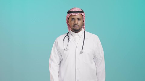 طلب المنع والتوقف عن فعل شيء ما, بورتريه مقرب لطبيب عربي خليجي سعودي يرتدي المعطف الطبي و السماعات و الشماغ ينظر الى الكاميرا و يشير بيده لطلب التوقف والابتعاد، خلفية خضراء