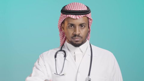 التوقف وعدم التقدم للأمام, بورتريه مقرب لطبيب عربي خليجي سعودي يرتدي المعطف الطبي و السماعات و الشماغ ينظر الى الكاميرا و يشير بيده لطلب التوقف والابتعاد، خلفية خضراء