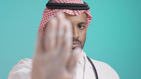 رفع اليد للحذر من الاقتراب, بورتريه مقرب لطبيب عربي خليجي سعودي يرتدي المعطف الطبي و السماعات و الشماغ ينظر الى الكاميرا و يشير بيده لطلب التوقف والابتعاد، خلفية خضراء