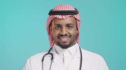 بورتريه مقرب لطبيب عربي خليجي سعودي يرتدي المعطف و السماعات الطبية و الشماغ، مفهوم الطب والرعاية الصحية، تقديم الخدمات الصحية، ايماءات وجه سعيد ويدين تدل على القبول والموافقة، خلفية خضراء