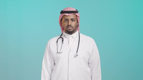 الفشل في تحقيق أمر ما، مفهوم التقييم السلبي والانزعاج، بورترية لطبيب عربي خليجي سعودي يرتدي المعطف و السماعة الطبية و الشماغ ينظر الى الكاميرا يشير بإصبع الابهام الى الاسفل بإيماءات الرفض وعدم القبول والرضا،خلفية خضراء