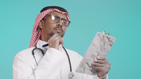 ايماءات التفكير فى شيء ما, العمل في القطاع الصحي في المملكة العربية السعودية,  تدوين الحالات المرضية ومتابعه المرضى, الاهتمام بالمرضى وتسجيل الملاحظات الطبيه، بورترية مقرب لطبيب عربي خليجي سعودي يرتدي المعطف و السماعة الطبية يقوم بكتابة تقرير طبي  , خلفية خضراء
