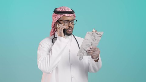 الرعاية الصحية باستخدام التقنية, تقديم الخدمات للمرضى بكل كفاءه, مفهوم الطب والرعاية الصحية, بورترية لطبيب عربي خليجي سعودي يرتدي المعطف الابيض و السماعات الطبية يتحدث بالهاتف المحمول  و يقوم بمراجعة الملاحظات، خلفية  خضراء