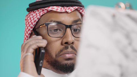 التواصل الطبي عن بعد, تقديم الخدمات للمرضى بكل كفاءه, مفهوم الطب والرعاية الصحية, بورترية مقرب لطبيب عربي خليجي سعودي يرتدي المعطف الابيض و السماعات الطبية يتحدث بالهاتف المحمول  و يقوم بمراجعة الملاحظات، خلفية  خضراء