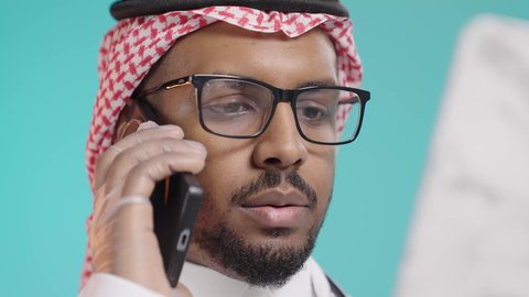Conducting remote medical consultations, providing services to patients with efficiency, the concept of medicine and healthcare, a close-up portrait of an Arab Gulf Saudi doctor wearing a white coat and medical headphones talking on a mobile phone and reviewing notes, green background.