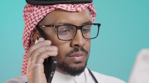 Providing services to patients with efficiency, the concept of medicine and healthcare, a close portrait of an Arab Gulf Saudi doctor wearing a white coat and medical headphones, talking on a mobile phone and reviewing notes, conducting remote medical consultations, green background.