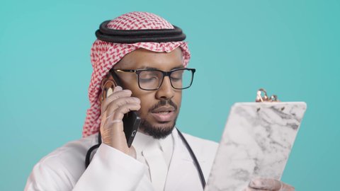 مفهوم الطب والرعاية الصحية, تقديم الخدمات للمرضى بكل كفاءه, بورترية مقرب لطبيب عربي خليجي سعودي يرتدي المعطف الابيض و السماعات الطبية يتحدث بالهاتف المحمول  و يقوم بمراجعة الملاحظات، اجراء الاستشارات الطبيه عن بعد، خلفية  خضراء