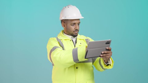 التنقل بين صفحات التواصل الاجتماعي، استخدام جهاز تقني حديث متطور، مفهوم الهندسة والإنشاء المعماري، مراجعة المخططات عبر الجهاز اللوحي، بورتريه لمهندس عربي خليجي سعودي يرتدي ستره و خوذة الحماية يمسك بيده جهاز التابليت المحمول، خلفية خضراء