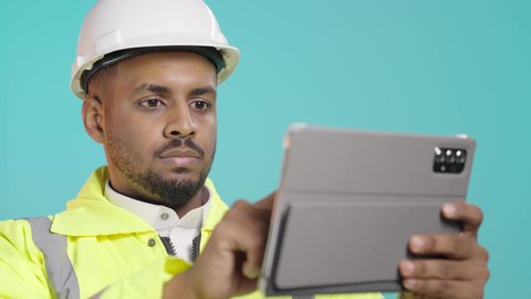 تصفح مواقع التواصل الاجتماعي، مفهوم الهندسة والإنشاء المعماري، مراجعة المخططات عبر الجهاز اللوحي، استخدام جهاز تقني حديث متطور، بورتريه مقرب لمهندس عربي خليجي سعودي يرتدي ستره و خوذة الحماية يمسك بيده جهاز التابليت المحمول، خلفية خضراء